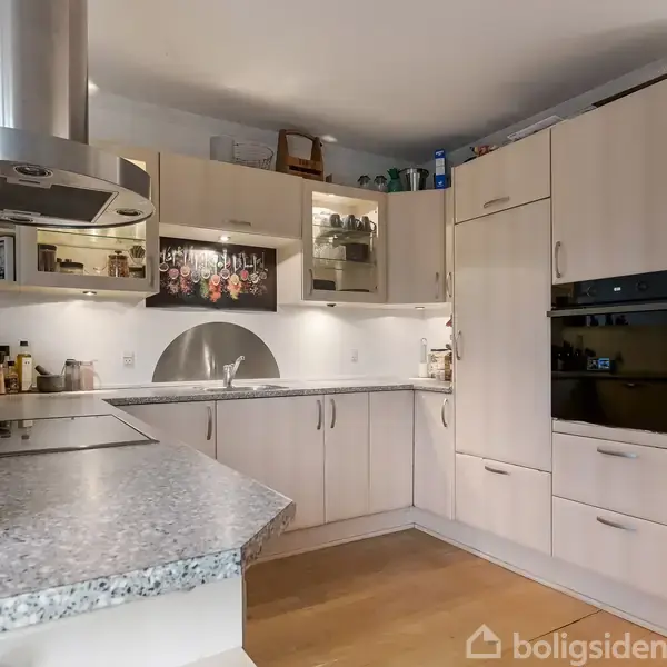 Et moderne køkken med lyse træskabe omgiver en hjørnevask. På væggen hænger et kunstværk, og der er indbygget ovn ved siden af skabe. Bordpladen er granitgrå.