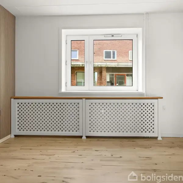 Et lyst rum har et vindue i midten, omkranset af en hvid radiator. Trægulvet komplementeres af en væg med trælister til venstre, og udenfor ses en murstensbygning.