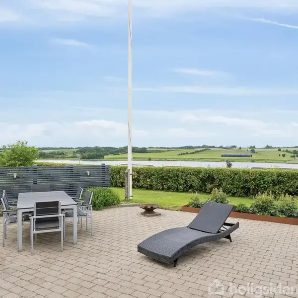 En lænestol og spisebord med stole står på en flisebelagt terrasse. Omkring er blomster i krukker, og landskab med grønne marker og buske ses i baggrunden under en blå himmel.