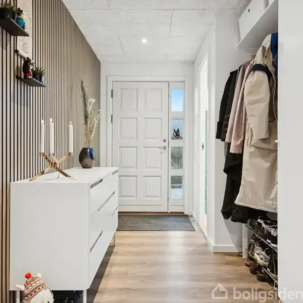 Garderobe står ved siden af en hvid kommode med lys og dekorationer, i en gang med trægulv. Døren i baggrunden slipper lys ind. Tøj hænger til højre.