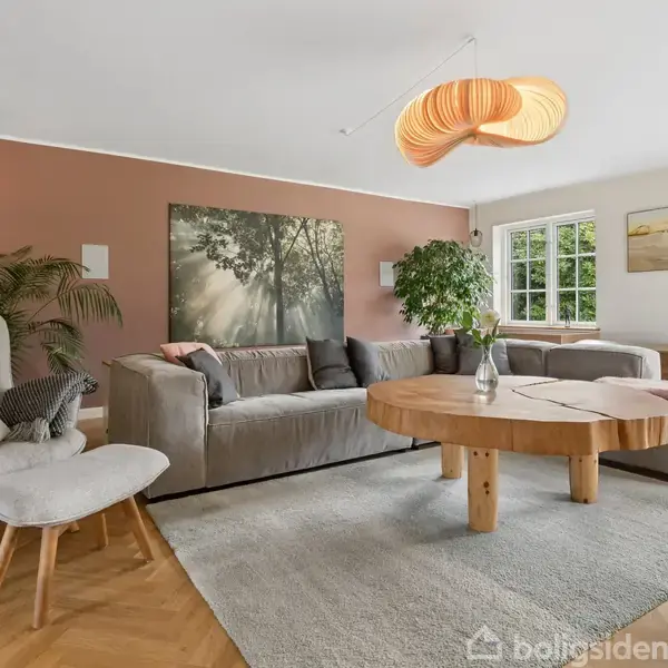En stor, grå sofa står i en stue med beige vægge. Et rundt træbord med en vase på står foran sofaen. Et stort vindue giver udsigt til en grøn have.