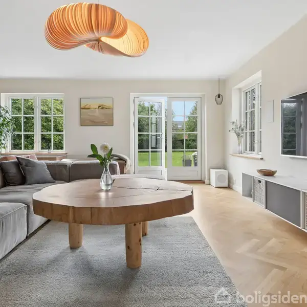 Stor, rund træbordplade med vase står på gulvtæppe. Omkranset af grå sofaer under kunstnerisk loftlampe. Fladskærms-tv på væggen, vinduer og glasdør åbner mod grøn have.