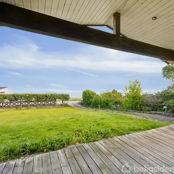 Verandaen har en trægulv, hvorfra man kan se en græsplæne, hækplanter og en sti. I baggrunden ses en klar blå himmel og et hus til venstre.