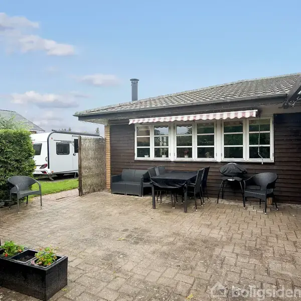 En terrasse med borde og stole står foran et hus med et stripet markise. Huset er omgivet af hæk og en parkeret campingvogn ses i baggrunden under en blå himmel.
