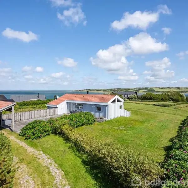 Hus ligger fredeligt omgivet af grønne buske og en græsplæne, med rødt tag og hvid facade. I baggrunden ses havet under en blå himmel med spredte skyer.