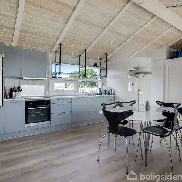 Et moderne køkken med grå skabe og indbygget ovn står til venstre. Et rundt spisebord med fire sorte stole er placeret midt i rummet. Rummet har en træloft og store vinduer med udsigt.