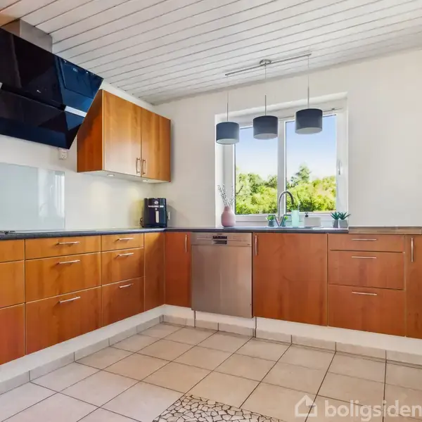 Køkken med træskabe og sort bordplade. En opvaskemaskine og kaffemaskine ses under og på bordpladen. Lyse fliser på gulvet. Et vindue med udsigt til grønne træer.