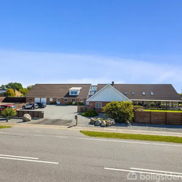 En stor moderne gårdbygning med brunlige tagsten står i midten, omgivet af parkeringsplads og have. Solrig dag med klar blå himmel og grønne træer i baggrunden.