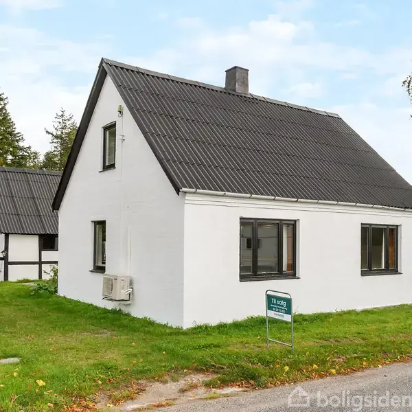 Et hvidt hus står på en grøn græsplæne med en til-salg-skilt foran. Det har et sort tag og en mindre bygning i baggrunden, omgivet af træer.
