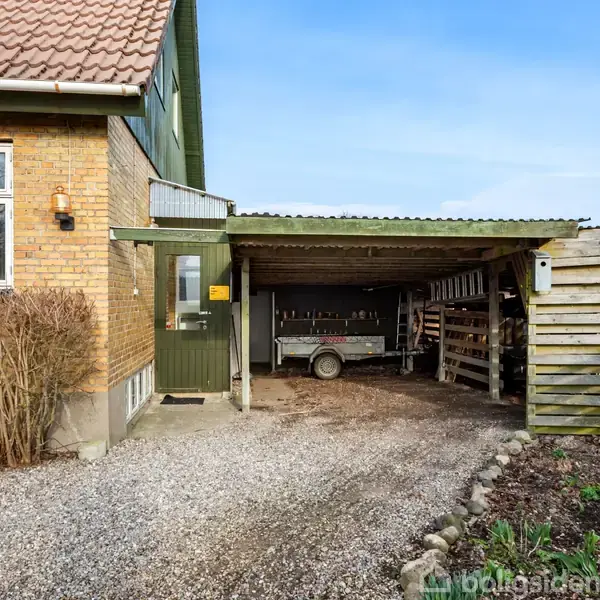 Hus med gult murstensfacade og grøn dør viser en carport med trailer parkering, omgivet af træhegn og grusvej, beliggende i et landligt område med marker i baggrunden.