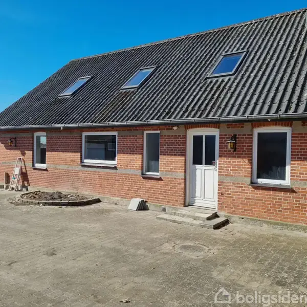 Rødstenshus med sort tegltag står på en stor flisebelagt gårdsplads. Tretten tagvinduer og to almindelige vinduer ses tydeligt. En stige læner mod huset nær en busk.