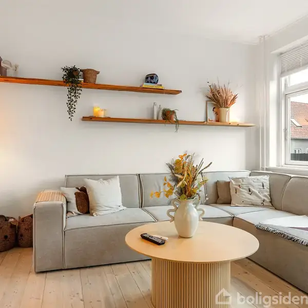 Grå sofa står i en stue med lyse træplanker på gulvet. Hylder bærer planter og dekorationer over sofaen. Rundt sofabord med vase. Vindue belyser rummet med dagslys.