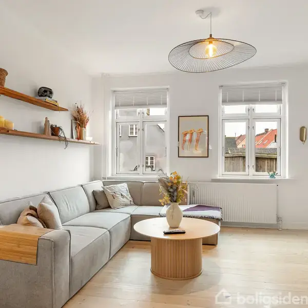 Gråt sofaarrangement står i stue med lyst trægulv. Sofaen er placeret under hylder med dekorationer. To vinduer giver udsigt til bygninger udenfor. Rundt sofabord i træ står centralt.