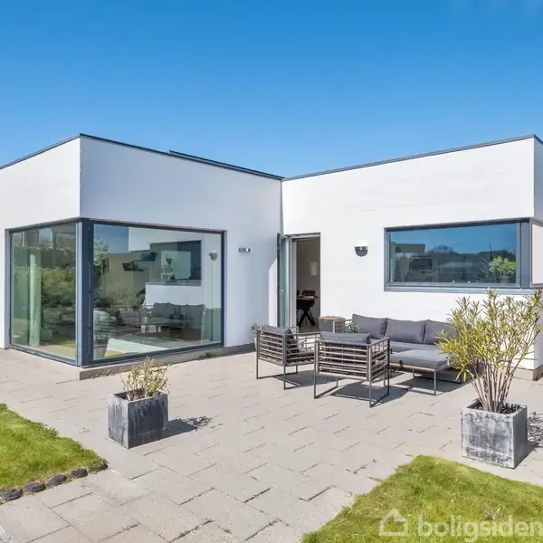 Moderne hvid villa med store glaspartier står på en solrig, flisebelagt terrasse. Omkranset af velplejet græsplæne og buske, rummer terrassen udendørsmøbler og dekorative planter i krukker.