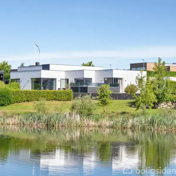 Et moderne, hvidt hus med fladt tag står stille ved en sø. Omgivet af grønne buske og træer, reflekteres huset i det klare vand.