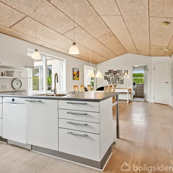 Køkkenø med hvidt skab, vask og opvaskemaskine; står centralt i lyst, moderne køkken-alrum med spisebord; omgivet af store vinduer og lyst trægulv.