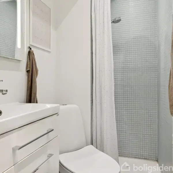 Et hvidt badeværelse med vask og skuffer til venstre, toilet i midten, og brusekabine med en hvid bruserforhæng til højre. Håndklæder hænger på væggen.