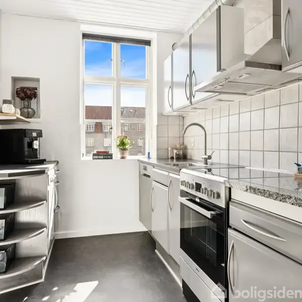 Køkken i moderne design med stålgrå skabe og hvide fliser over granitbordpladen. Komfur og opvaskemaskine langs væggen. Enkelt vindue giver udsigt til nabobygninger og dagslys ind.