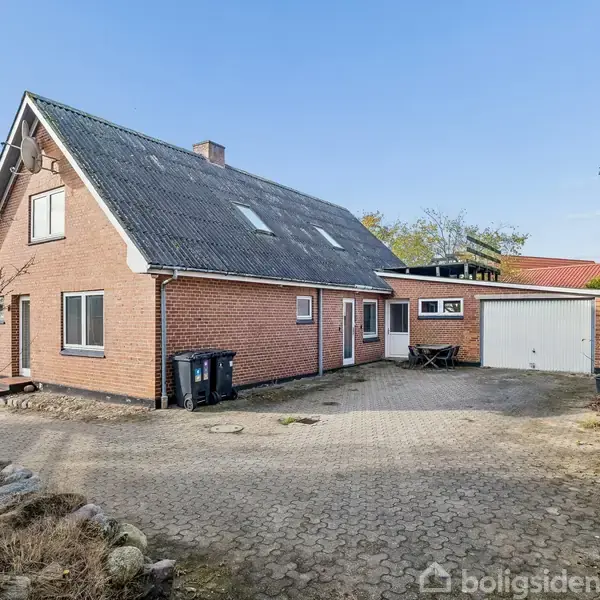 Et rødt murstenshus med sort tag står stille på en flisebelagt gårdsplads. Omgivet af træer og en garagebygning i et boligområde.