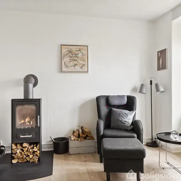 En moderne brændeovn med ild står til venstre, omgivet af brændestabler. En sort lænestol med fodskammel står ved siden af et lille bord og en gulvlampe. Væggene er lyse med kunstværker, og der er et vindue med udsigt.