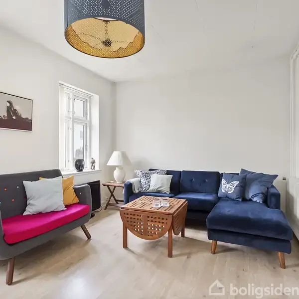 En stue med to sofaer, en grå med pink sæde og en blå med puder, står omkring et træsofabord. En lampe hænger ovenover, og et vindue i hjørnet giver lys.