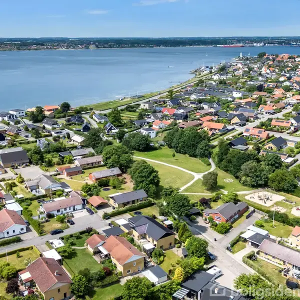 En række huse med røde og sorte tage ligger tæt på hinanden, omgivet af veje og grønne områder. En park med stier og træer er centralt placeret, tæt ved en bred flod eller sø.