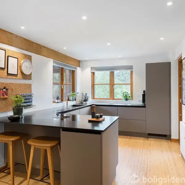 Moderne køkken med mørkegrå køkkenø omgivet af træstole, står i et rum med lyse trægulve og træbjælker. Vinduerne giver naturligt lys, mens en opslagstavle hænger på væggen.