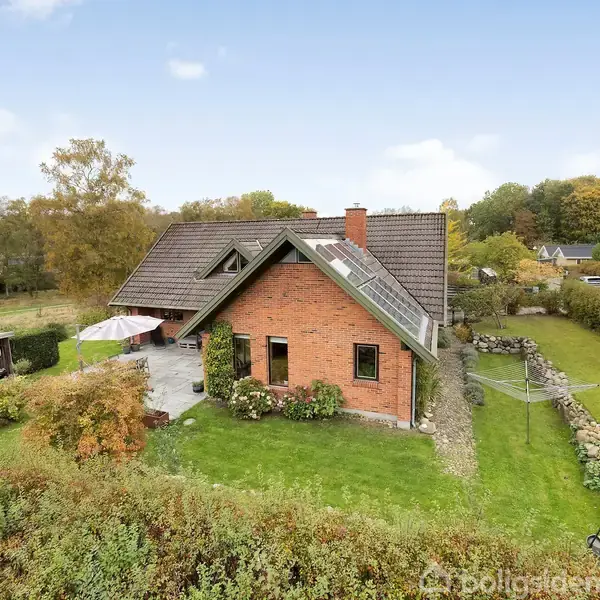 Et rødt murstenshus står i en grøn have, omgivet af hække og træer. En terrasse med parasol ligger ved siden af huset. Boligområde i baggrunden.