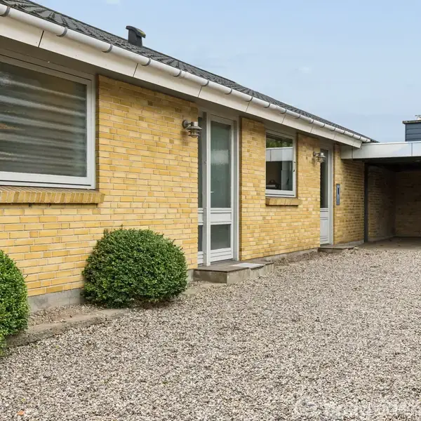 Gul murstensrækkehus står stille på en grusbelagt gårdsplads. En dør og vinduer pryder facaden, mens en overdækket carport ses til højre. To klippede buske flankerer stien.