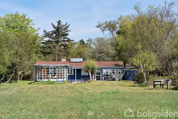 Et lille, blåt sommerhus med rødt tag står stille omgivet af grønne træer og buske. Foran huset er en velplejet græsplæne med en træbænk til højre.