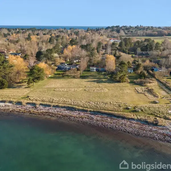 Kystlinje strækker sig langs klart vand, omgivet af frodig, grøn natur og spredte træer. I baggrunden ses enkelte huse indlejret i skovområde under en klar, blå himmel.