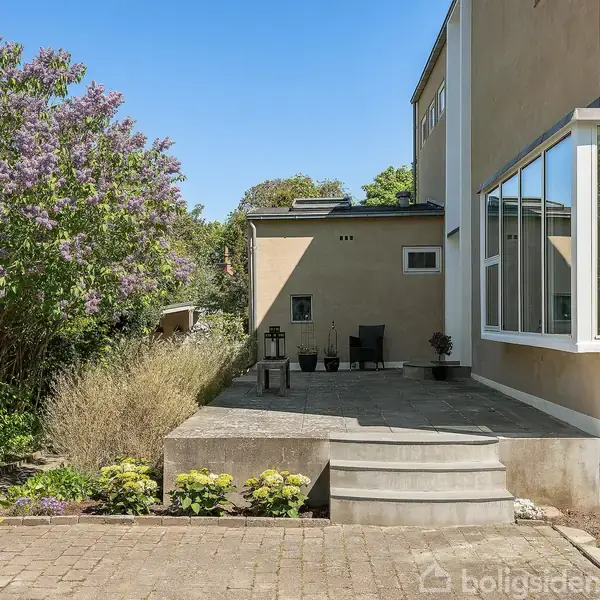 Moderne hus; terrasse med runde trappetrin; omgivet af blomstrende buske og grønne træer; klart lys baggrund.