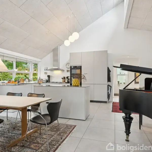 Flygel står i en moderne, lys stue-køkken kombination med højt til loftet. Køkkenet har et spisebord med stole, og store vinduer bringer lys ind i rummet.