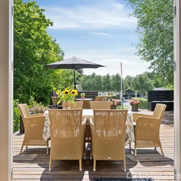 En udendørs træterrasse med et bord omgivet af flettede stole. Bordet er pyntet med solsikker i en vase. Parasollen står åben, omgivet af frodige grønne træer og en klar himmel.