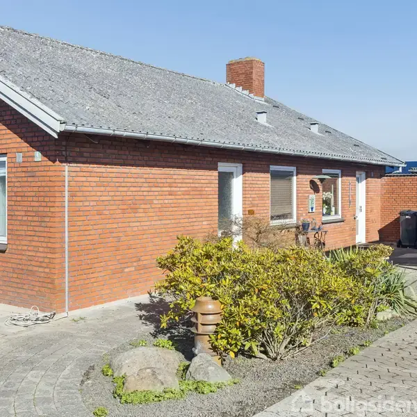 Rødt murstenshus med stigende tag og skorsten. Huset har flere vinduer. Foran er der en have med buske og en stenbelagt gangsti. Carport til højre.