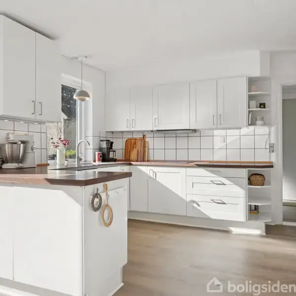 Køkken med hvide skabe og træbordplader. En køkkenmaskine står på bordet. Lyse klinker pryder væggen, mens et vindue tilføjer naturligt lys. Gulvet er lyst træ.