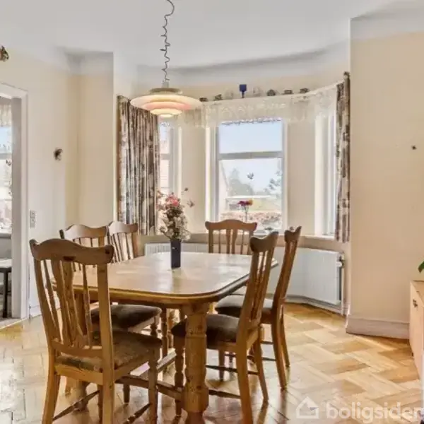 Et spisebord med seks træstole står under en pendellampe i et lyst rum med store vinduer. En vase med blomster pryder bordet. I baggrunden ses en stue.