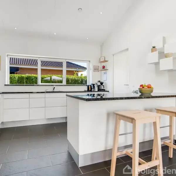 Et lyst køkken med hvide skabe omkranser et sort bordplade. To træstole står ved en køkkenø. Store vinduer giver udsigt til en grøn have. Ingen tekst er synlig.