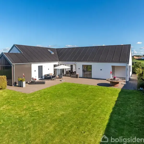 En moderne, hvid villa med sort tag står stille i en velplejet have med grøn græsplæne og terrasse. Hække omgiver området under en klar blå himmel.