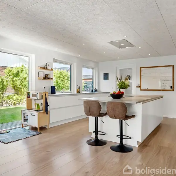 Køkken med en stor ø, to barstole og planter, indrammes af hvide vægge. Store vinduer giver udsigt til en grøn have. Rengjort og moderne design. Spilleområde for børn til venstre.