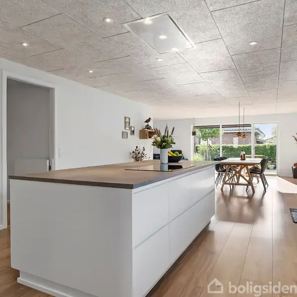 Hvid køkkenø med planter står i et moderne køkken-alrum. Til venstre er en indbygget ovn. I baggrunden er et spisebord ved vinduer, der åbner ud til en have.