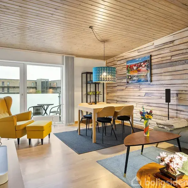 Gult lænestol står ved siden af en sofa i en stue med trægulv og træloft. Et spisebord med stole står i nærheden, og en balkon ses udenfor glasdøren.