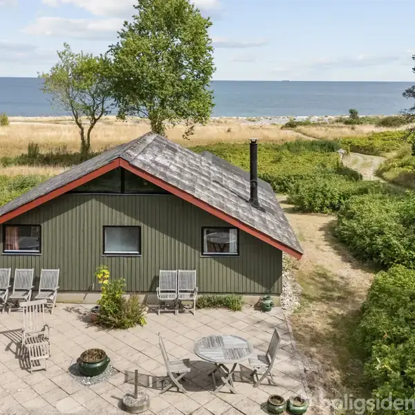 Et grønt sommerhus med sort tag står ved en terrasse med havemøbler. Baggrunden viser en strand og havet, omgivet af grønne buske og træer.
