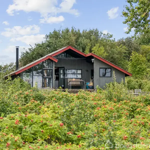 Et gråsort træhus med rød tagkant står i en frodig have fyldt med grønne buske og røde blomster, omgivet af træer under en blå himmel med spredte skyer.