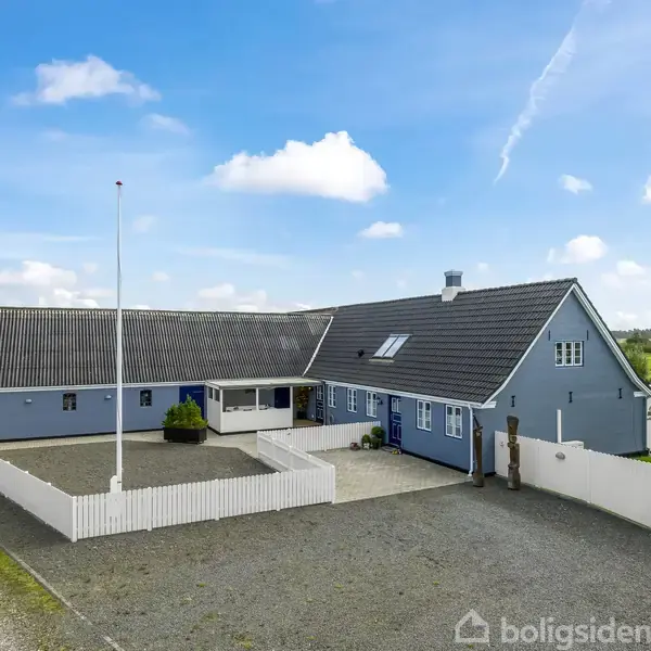 Blåt hus med mørkt tag står stille omgivet af hvidt stakit og grusplads. I baggrunden ses grønne marker og en klar blå himmel med få skyer.