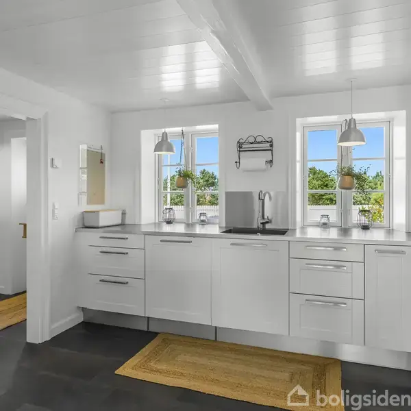 Et køkken med hvide skabe og skuffer står i et lyst rum. To vinduer over køkkenbordet giver udsigt til grønne træer udenfor. To tæpper dækker gulvet.