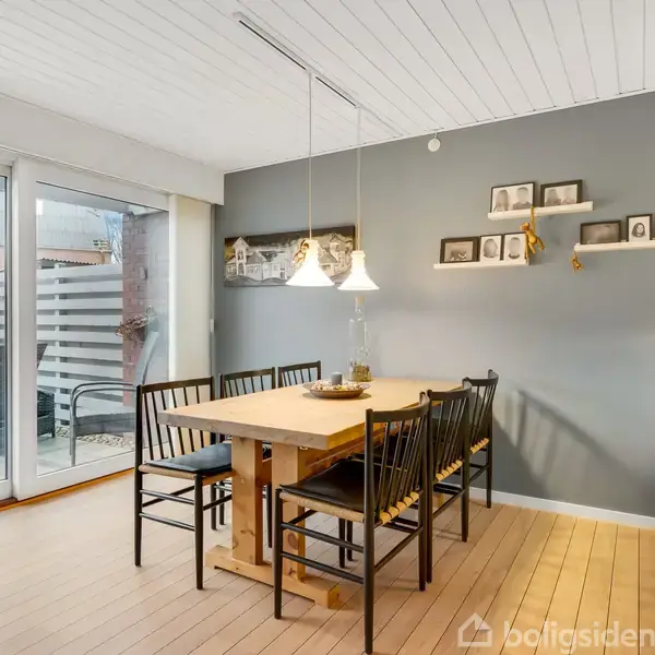 Et spisebord med sorte stole står i et lyst spisestueområde. Væggene har billedrammer, og store glasskydedøre fører ud til en terrasse med møbler.