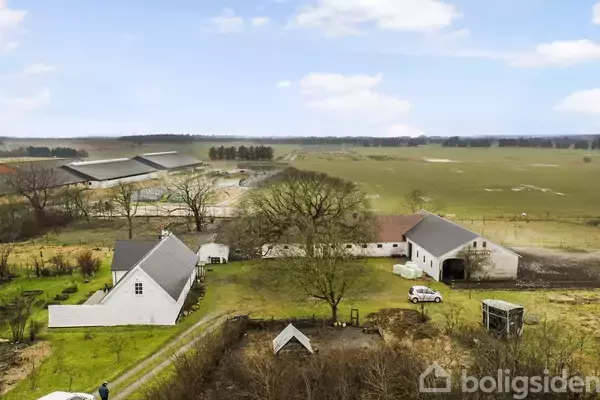 Gårdbygninger omgivet af åbne marker, med flere hvide bygninger og en parkeret bil i gårdspladsen; landskabet har træer og en vid udsigt over markerne under en blå himmel.