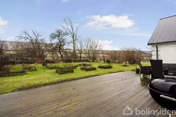 Terrassen har træplanker og rummer sorte møbler. I baggrunden findes en have med græs og højbede, omkranset af træer, under en lys himmel.
