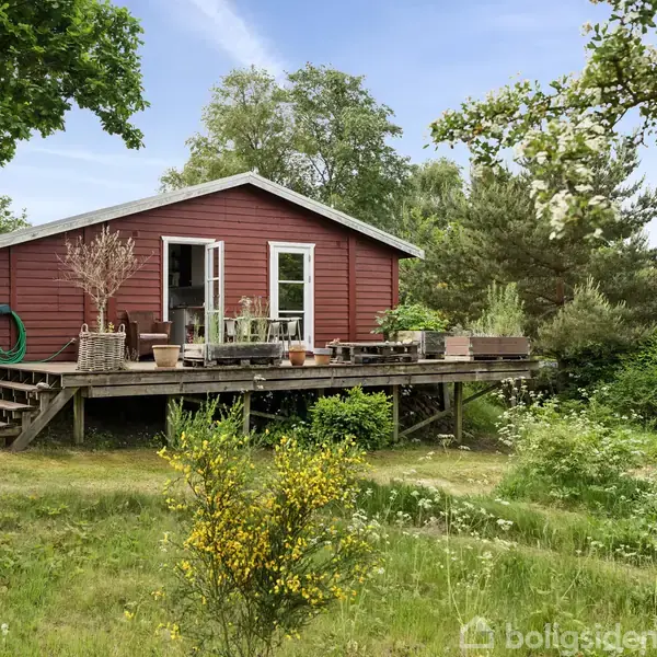Rødt træhus står stille på en græsplæne omgivet af frodig vegetation. En træterrasse med blomsterkrukker og havemøbler flankerer huset, mens grønne træer omkranser området.